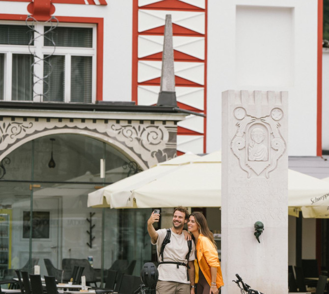 Two cyclists stand in front of a building decorated in red and white, posing for a photo with a mobile phone, their bikes beside them.