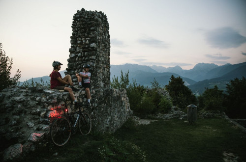 Two cyclists taking a break by an old castle wall, surrounded by an evening backdrop of a mountainous landscape.