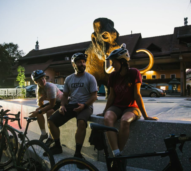Three cyclists are sitting by the mammoth statue in the square, illuminated by soft light with the cityscape in the background.