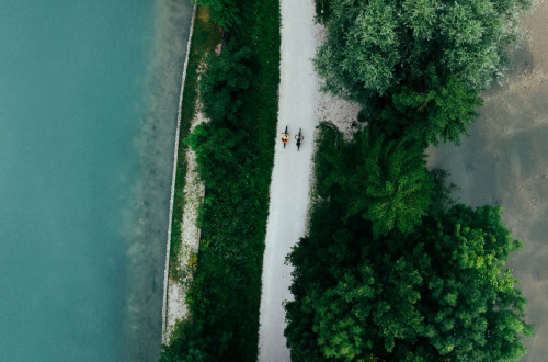 An aerial shot of cyclists on a white gravel path beside a turquoise-blue river and lush vegetation.
