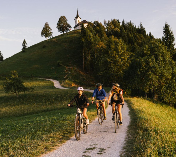 A family is cycling along a path in the middle of a meadow, with a small hill and a church at the top in the background.