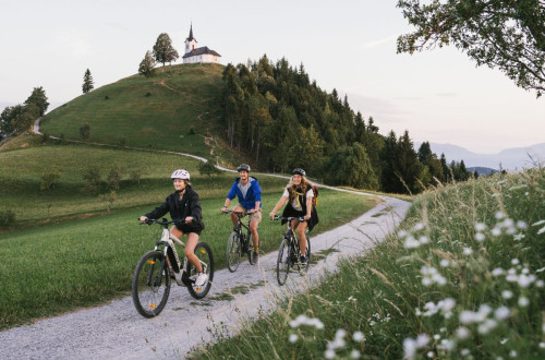 A family is cycling along a grassy path, with a small hill and a church on top in the background.