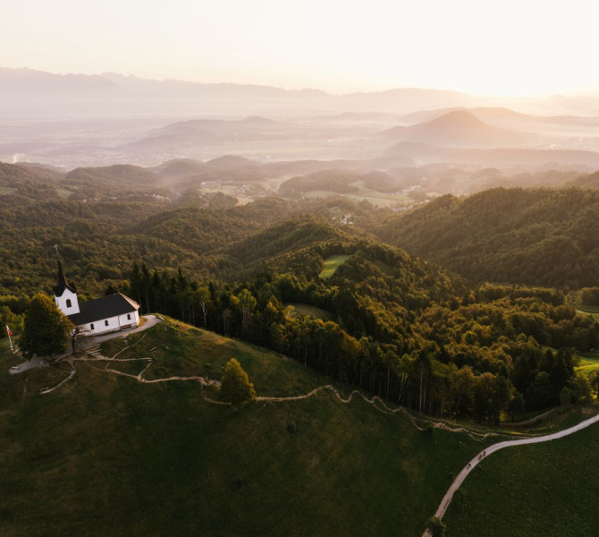 A view from above of a small church on top of a hill, with a backdrop of wooded hills.