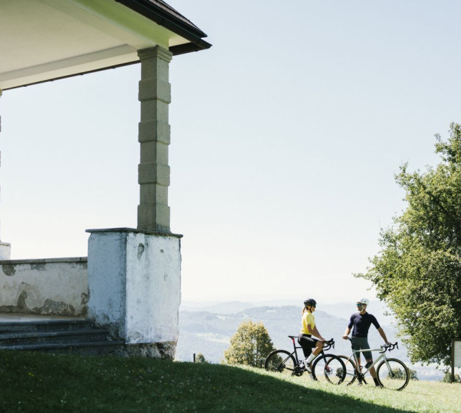 Two cyclists stand beside their bicycles on a meadow in front of a vintage pavilion, with mountains and lush green landscapes in the background.