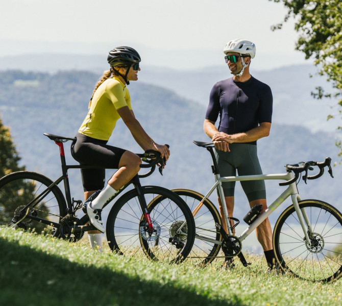 A male and female cyclist stand beside their bicycles on a meadow, dressed in sporty cycling attire, engaged in conversation, with mountain views in the background.