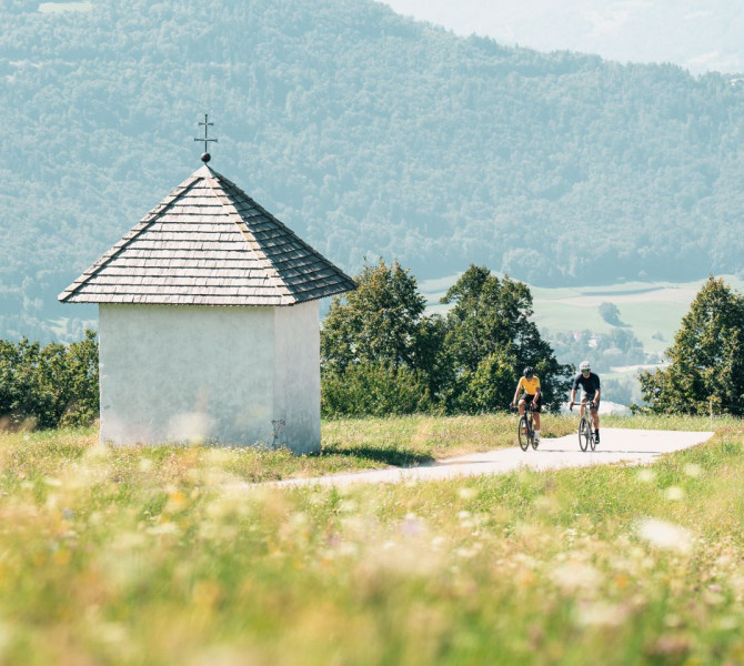 A male and female cyclist ride past a small chapel on a green meadow, with hills in the background.