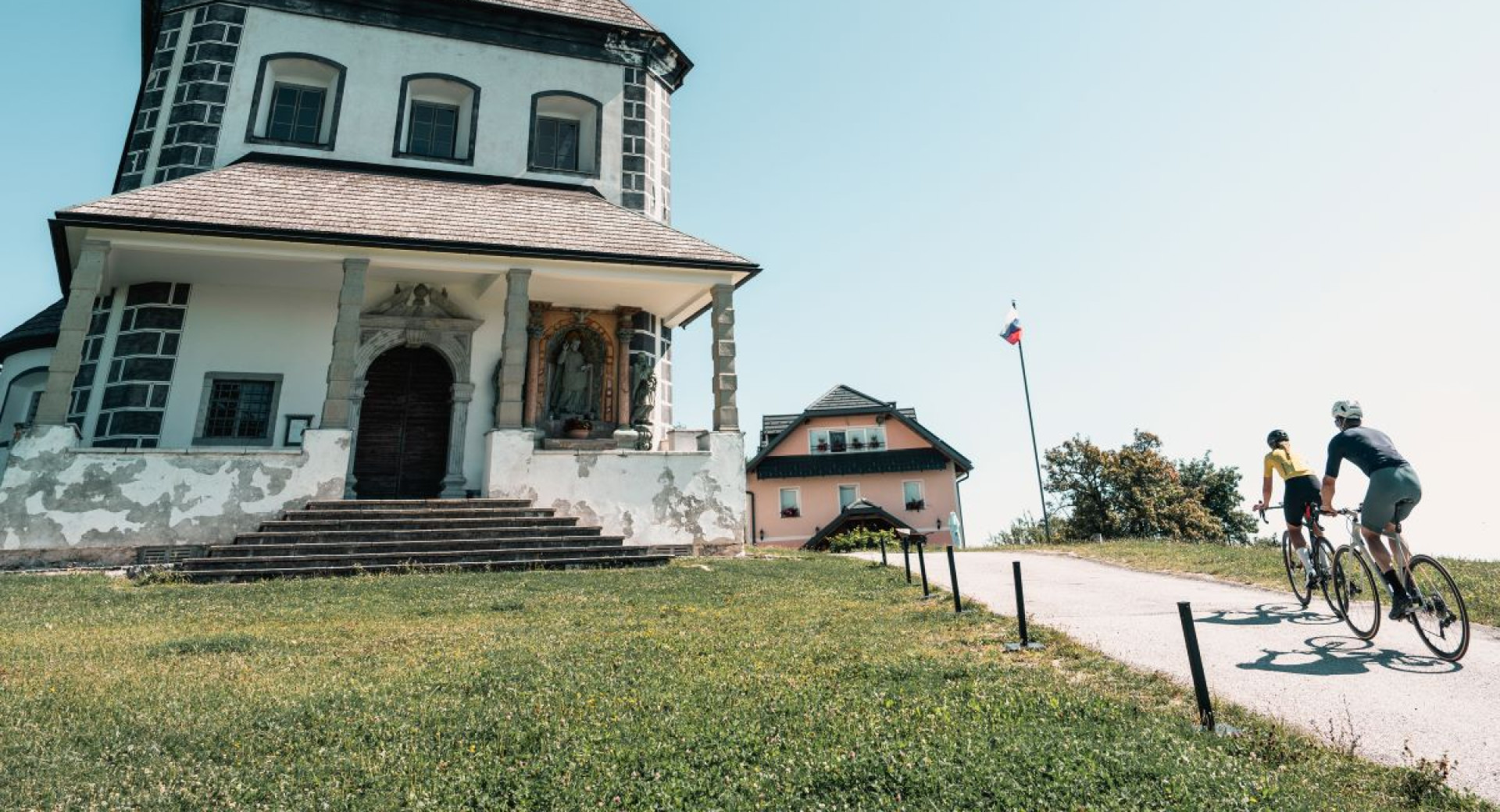 Kolesar in kolesarka se peljeta po poti proti stavbi z zgodovinsko fasado in slovenskimi zastavami, z zelenim travnikom in modrim nebom v ozadju.