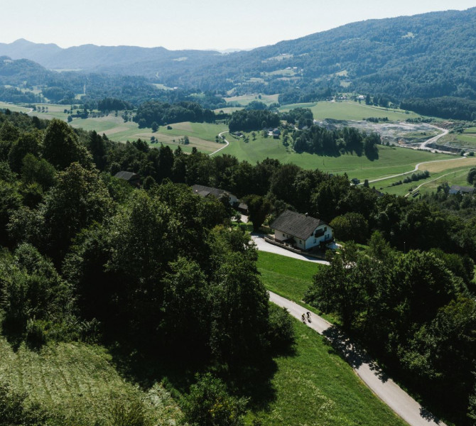 Two cyclists ride along a path through a green landscape, surrounded by hills and pastures, with distant mountains in the background.