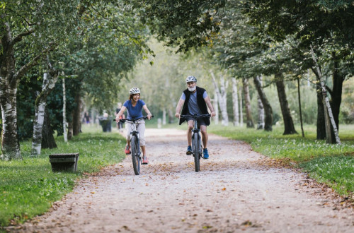 Two cyclists, a man and a woman, riding along a path lined with trees, surrounded by lush green countryside, chatting and smiling.