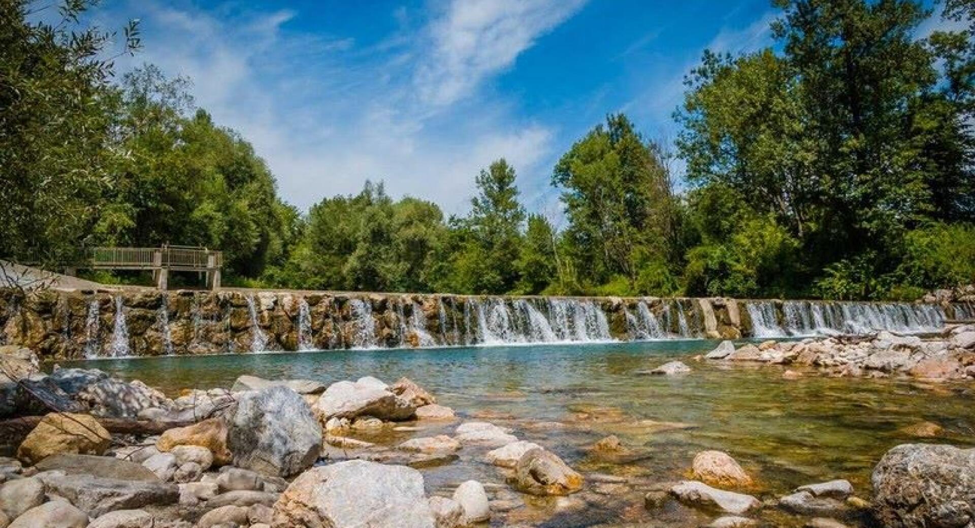 Cista reka s slapovi tece mimo skalnatega obmocja, obdana z gozdom in modrim nebom.