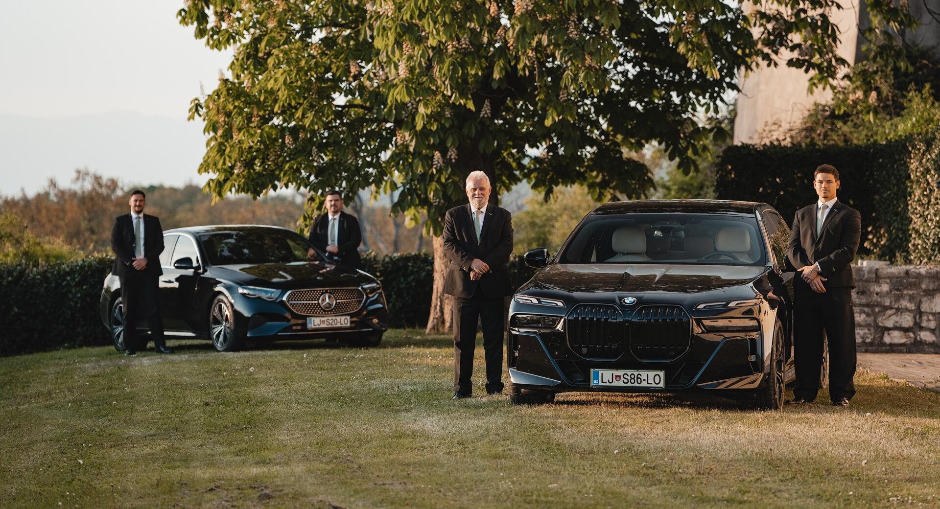Drivers in black suits standing next to elegant black cars on the grass.
