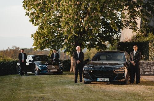Drivers in black suits standing next to elegant black cars on the grass.