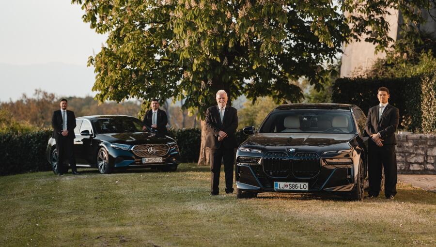 Drivers in black suits standing next to elegant black cars on the grass.