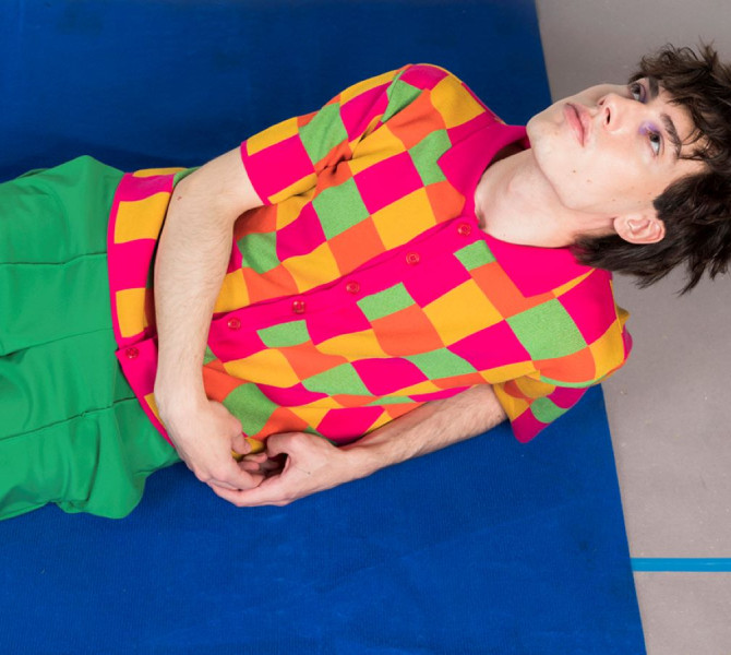 A man posing lying on the floor, dressed in green pants and checkered green, pink and orange shirt.