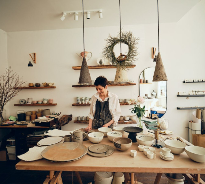 A ceramics studio with a smiling creator behind a wooden table, surrounded by dishes and decorations.