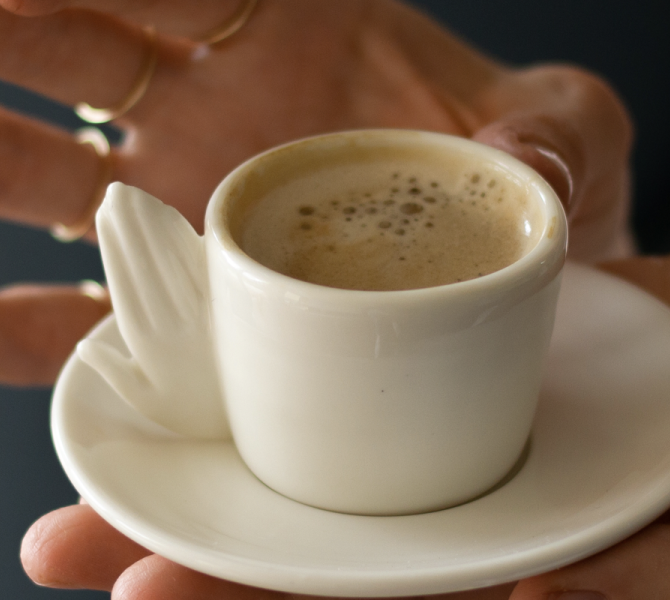 A white cup of coffee with foam on a saucer, held in hands with gold rings.
