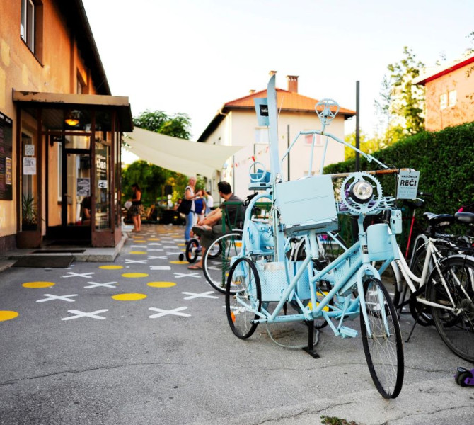 A vibrant outdoor space with a colorful bike installation and people socializing, walking, and enjoying the setting near a community building, with playful painted designs on the ground.