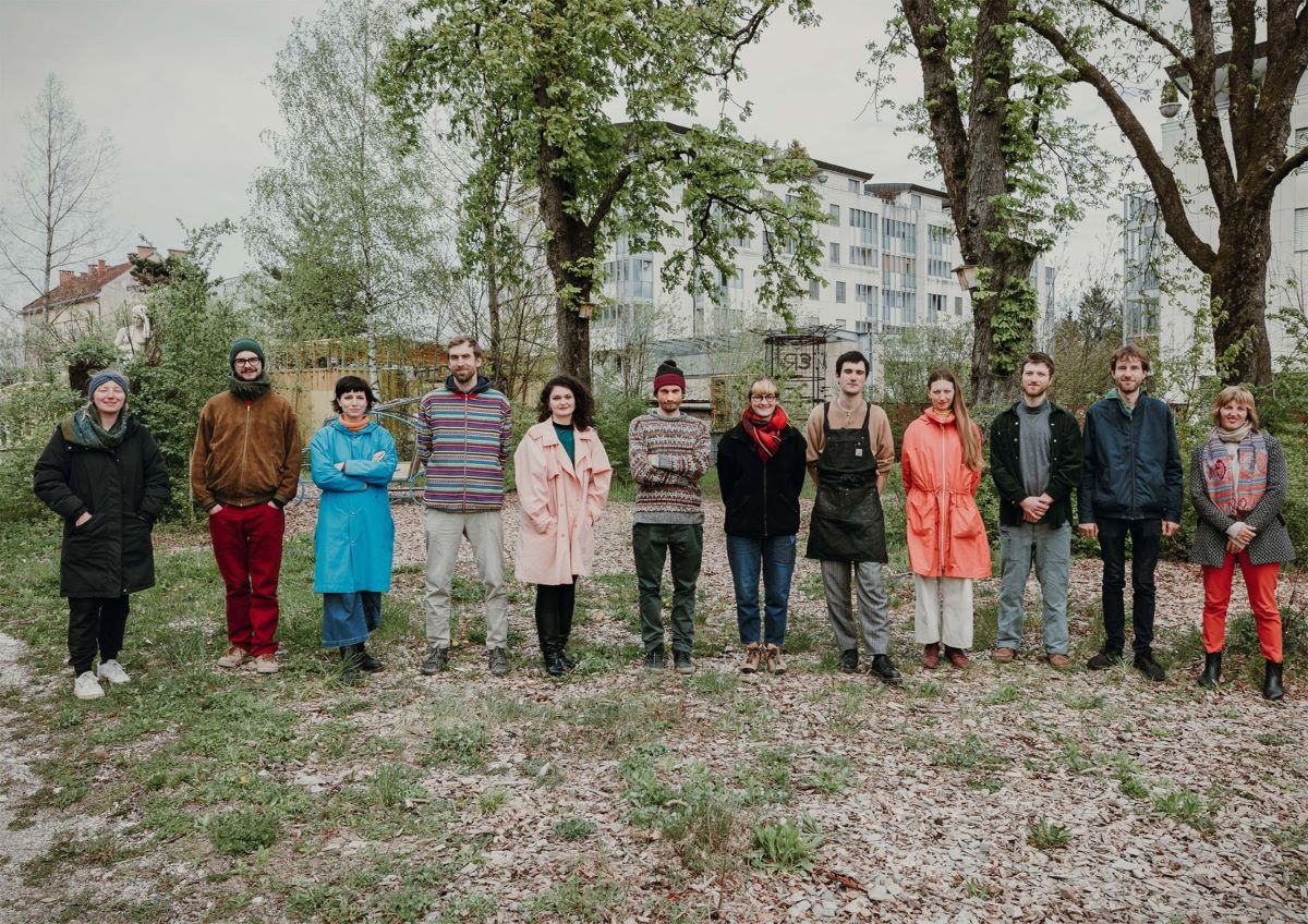 A group photo of thirteen people standing in a row outdoors in a green city park, surrounded by trees and fallen leaves, dressed in colourful clothing suitable for cold weather.