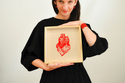 Donna in abito nero che tiene un'opera d'arte incorniciata con un'immagine rossa, sorridente e con dettagli di gioielli rossi.