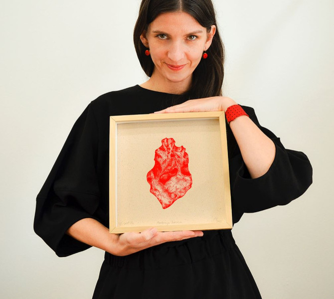 A woman in a black dress holding a framed artwork with a red image, smiling and wearing red jewelry details.