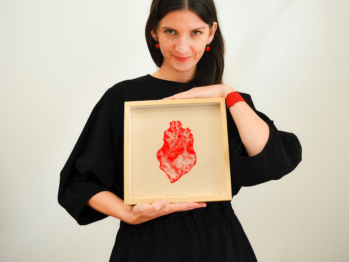 Donna in abito nero che tiene un'opera d'arte incorniciata con un'immagine rossa, sorridente e con dettagli di gioielli rossi.
