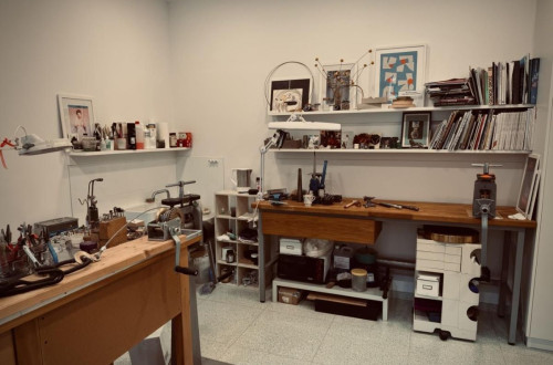 Werkstattbereich mit Werkzeugen und Materialien zur Schmuckherstellung, mit einem Arbeitstisch und Regalen, die verschiedene Gegenstände und Literatur enthalten.