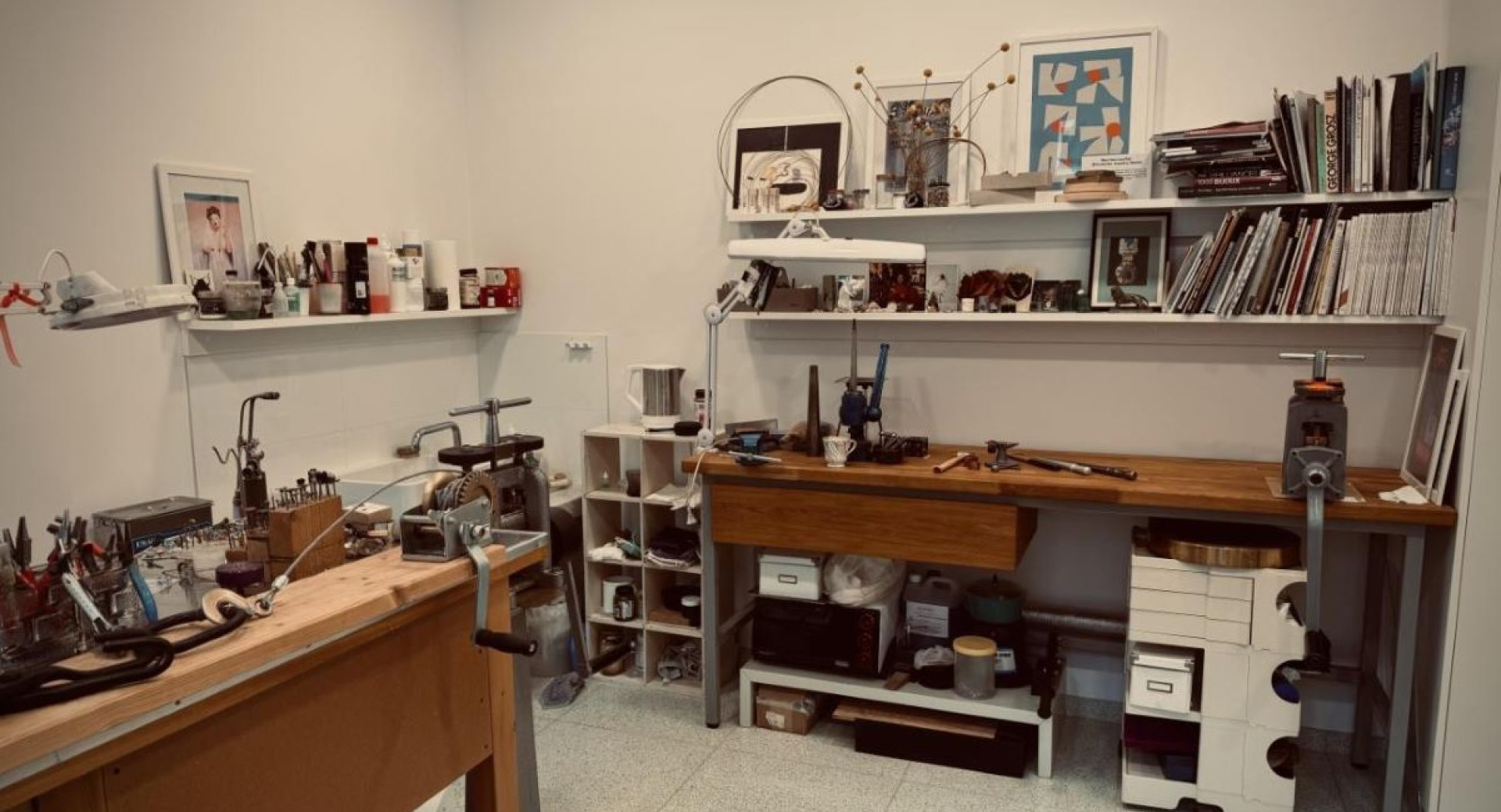 Werkstattbereich mit Werkzeugen und Materialien zur Schmuckherstellung, mit einem Arbeitstisch und Regalen, die verschiedene Gegenstände und Literatur enthalten.