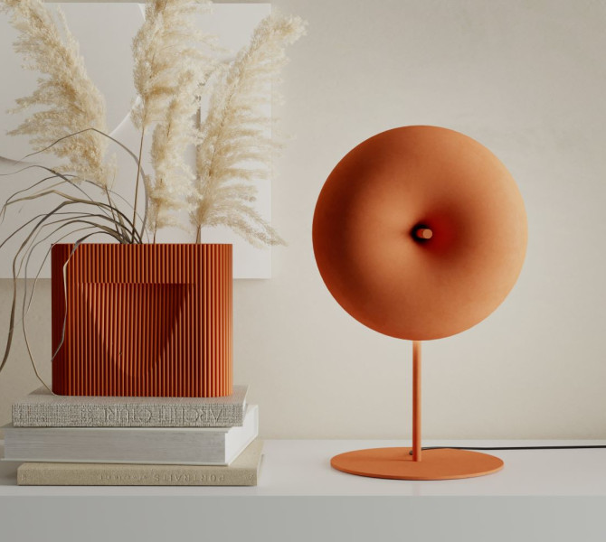 A contemporary minimalist interior featuring a orange-colored designer lamp with an round shape, a ribbed dark orange vase with pampas grass, and a stack of books on a sleek white surface.