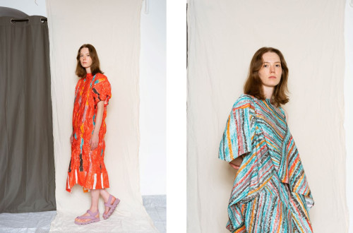 A woman in a loose, colorful striped dress with a draped silhouette, standing against a neutral fabric backdrop.