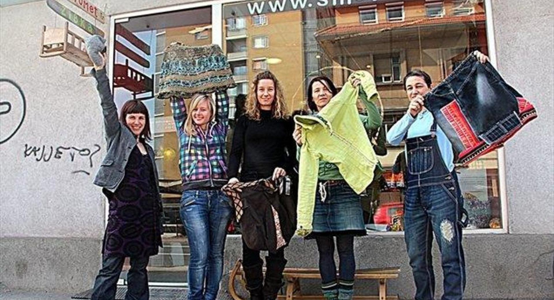 Un gruppo di cinque persone si trova davanti al negozio "Smetumet," mostrando abiti e accessori riciclati.