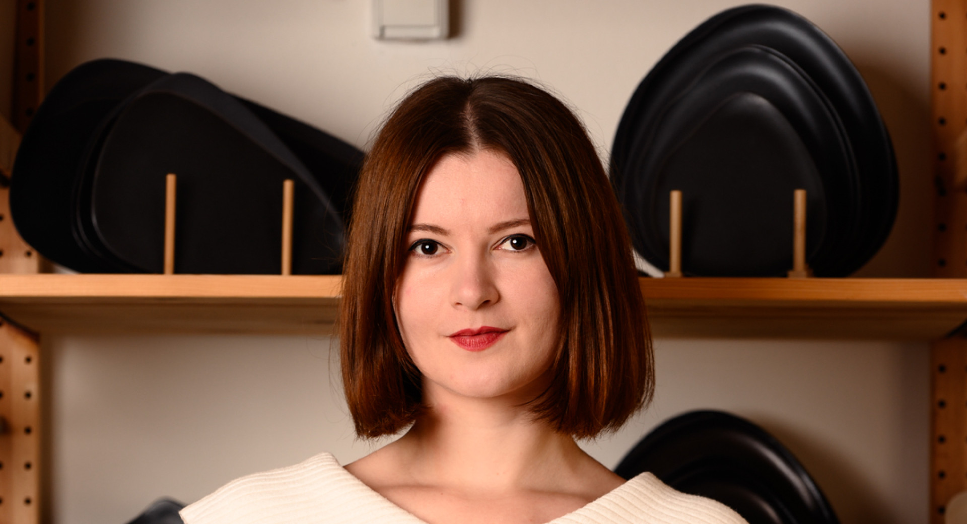 A woman standing in a ceramic studio, holding a black and a white handmade bowl, surrounded by pottery pieces.