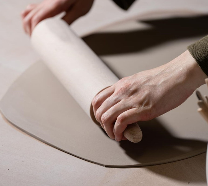 A close-up of hands rolling out clay with a rolling pin, with ceramic tools in a cup nearby.