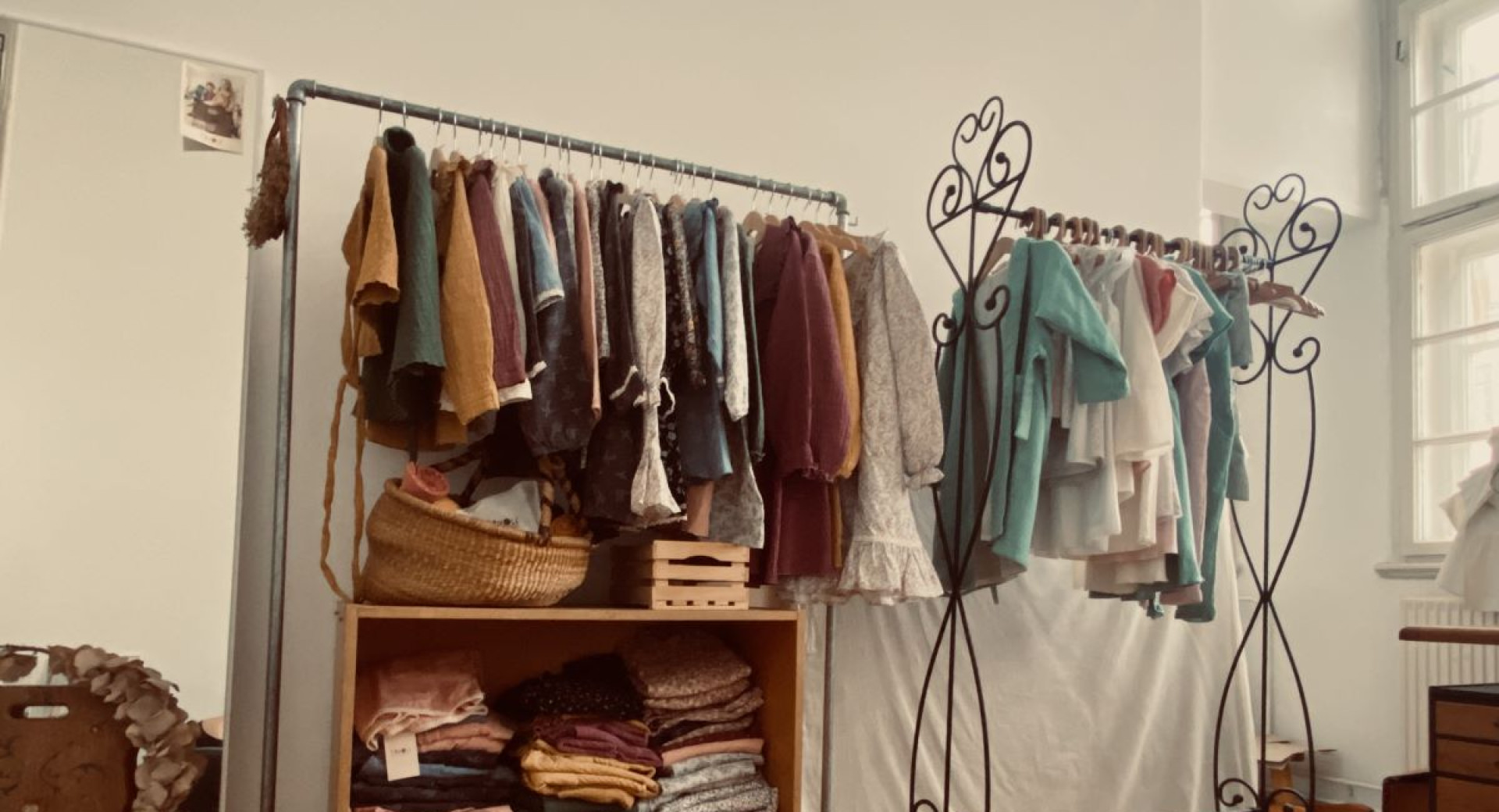A boutique or showroom displaying handmade, vintage-style clothing on metal racks, with neatly folded textiles on wooden shelves.