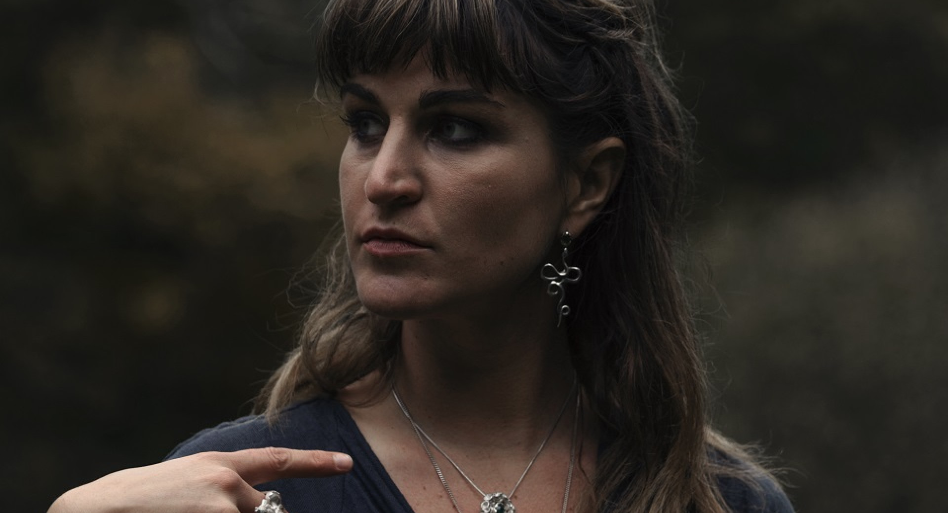 A woman with brown hair, wearing necklaces, rings, and earrings.