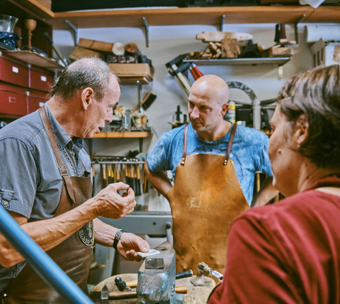 Zwei Teilnehmende hören einem Mentor in einer Goldschmiedewerkstatt aufmerksam zu, der ihnen am Arbeitstisch den Metallbearbeitungsprozess erklärt.