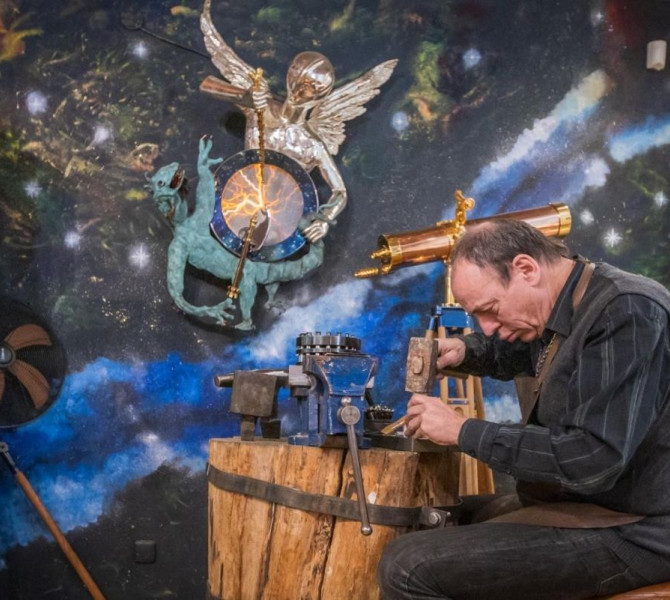 A goldsmith working on a jewelry piece at a wooden workbench, in a shop decorated with celestial and artistic elements.