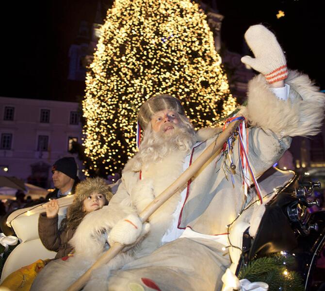A man dressed as Dedek Mraz sits on a carriage with a child, with a large Christmas tree illuminated in the background.