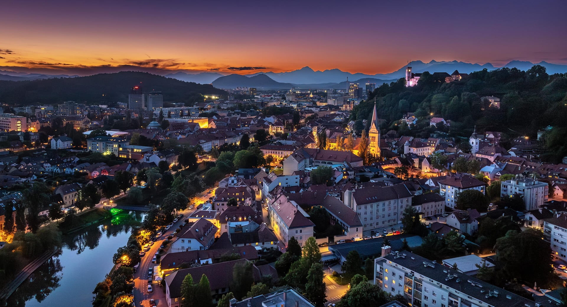 View of the city centre at sunset, with a river running through it, illuminated streets, a castle on the hill, and mountains in the background.