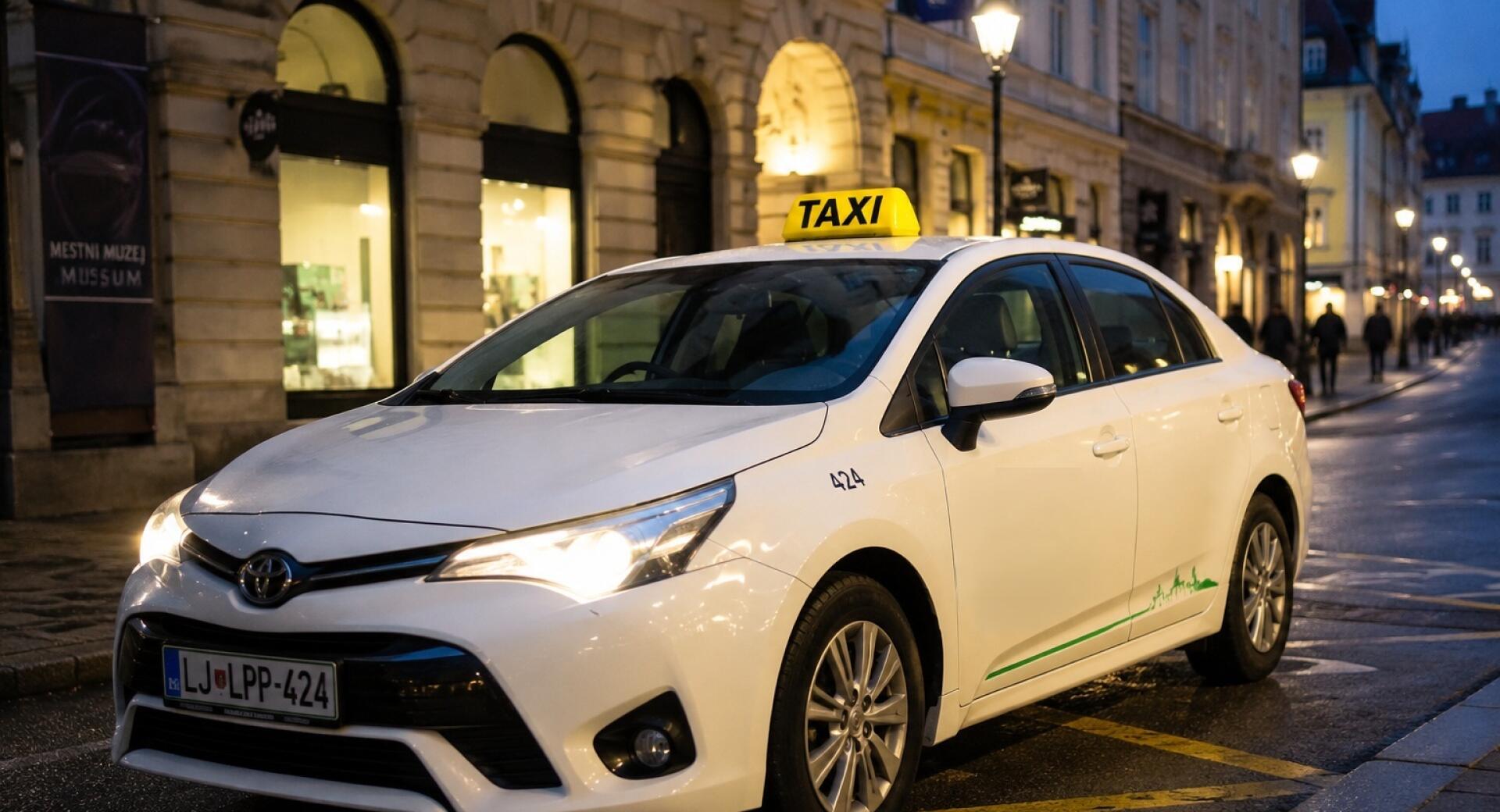 Un véhicule de taxi blanc avec un chauffeur et un panneau 'TAXI' jaune sur le toit se tient dans une rue de la ville éclairée par des lumières du soir. À l'arrière-plan, les fenêtres des magasins sont illuminées.