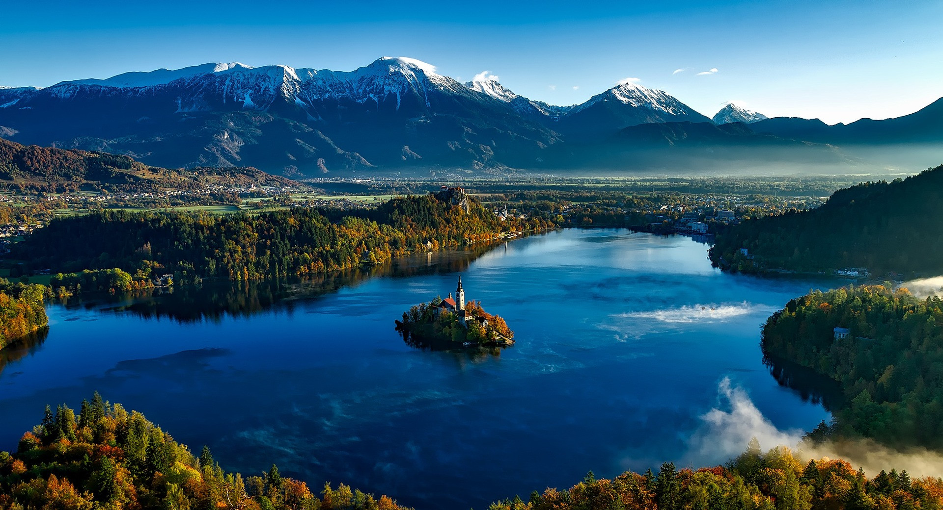 Panoramablick auf den Bleder See mit der Insel und der Kirche in der Mitte, im Hintergrund erheben sich schneebedeckte Berge.