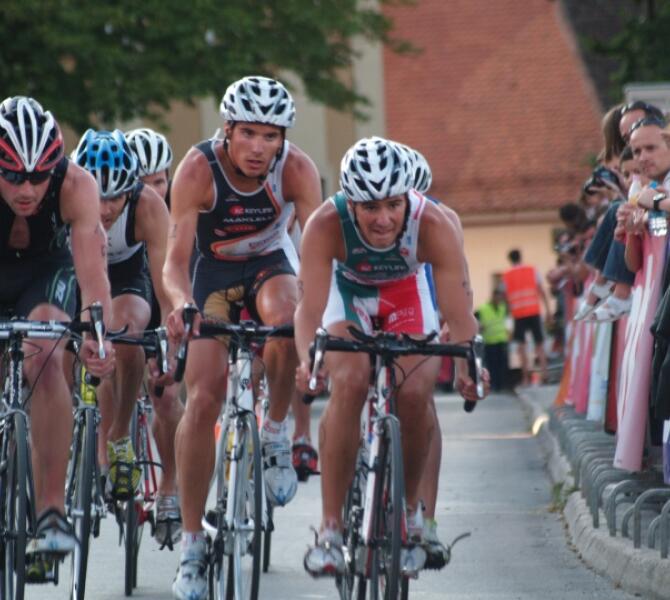 Cyclists in a close race on a street, with onlookers cheering them on from the sides.