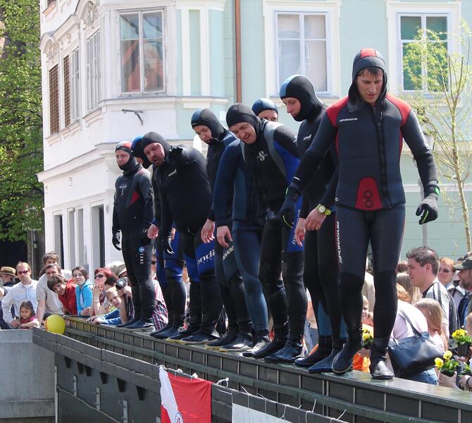Skupina potapljačev v neoprenskih oblekah se pripravlja na skok v reko, obkroženi z občinstvom v mestnem okolju.