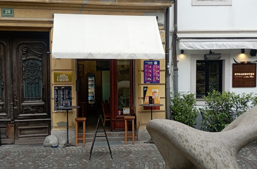 Entrée du bar avec un auvent blanc, des tables hautes noires et des tabourets de bar en bois.