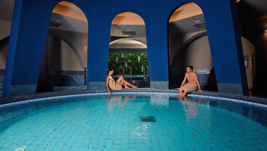 A man and a woman are sitting by a large circular pool, dipping their feet in the water.