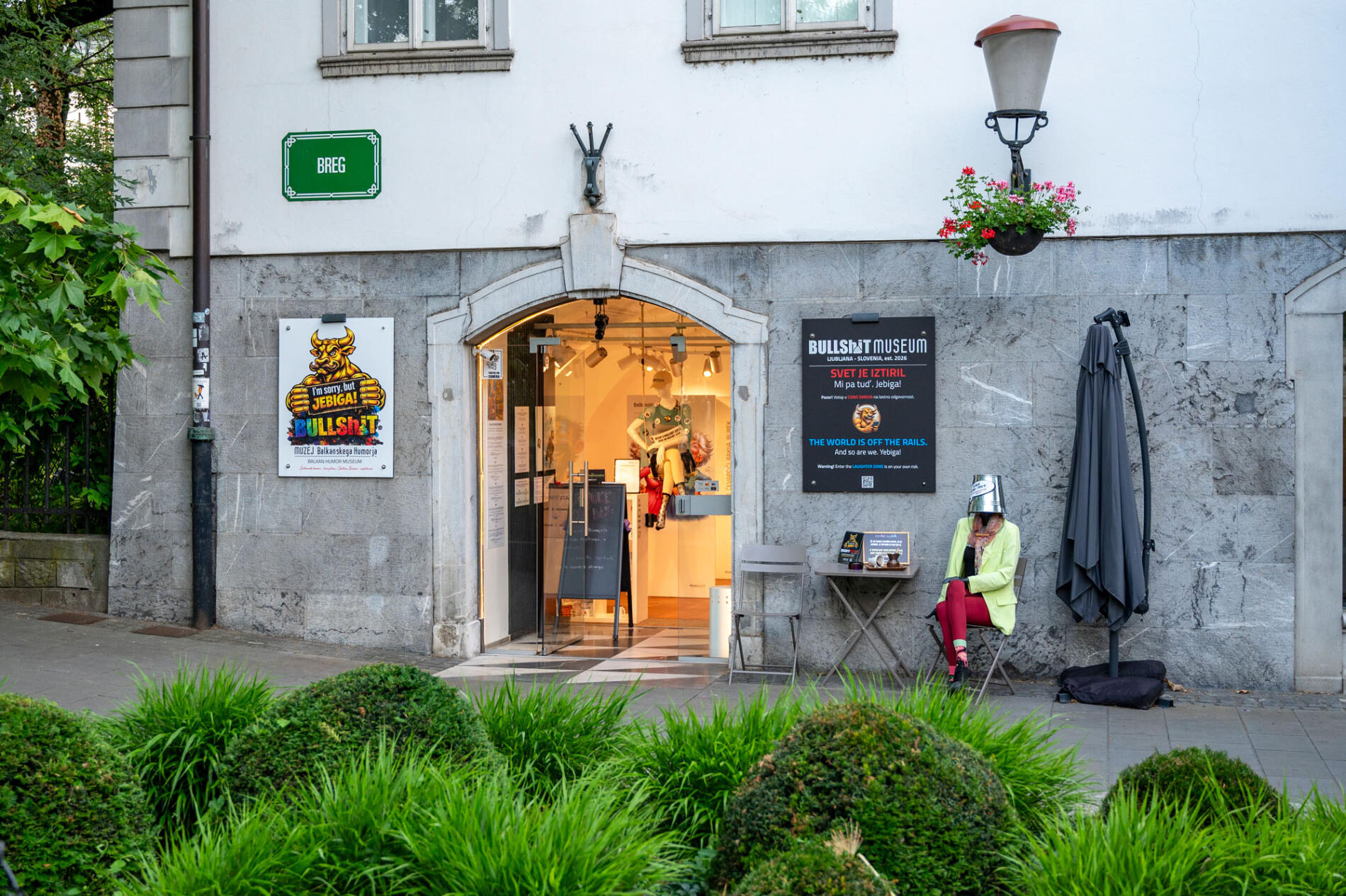 Vhod v muzej v Ljubljani, ob katerem sedi nenavadno oblecena figura, medtem ko v ospredju raste urejeno zelenje.