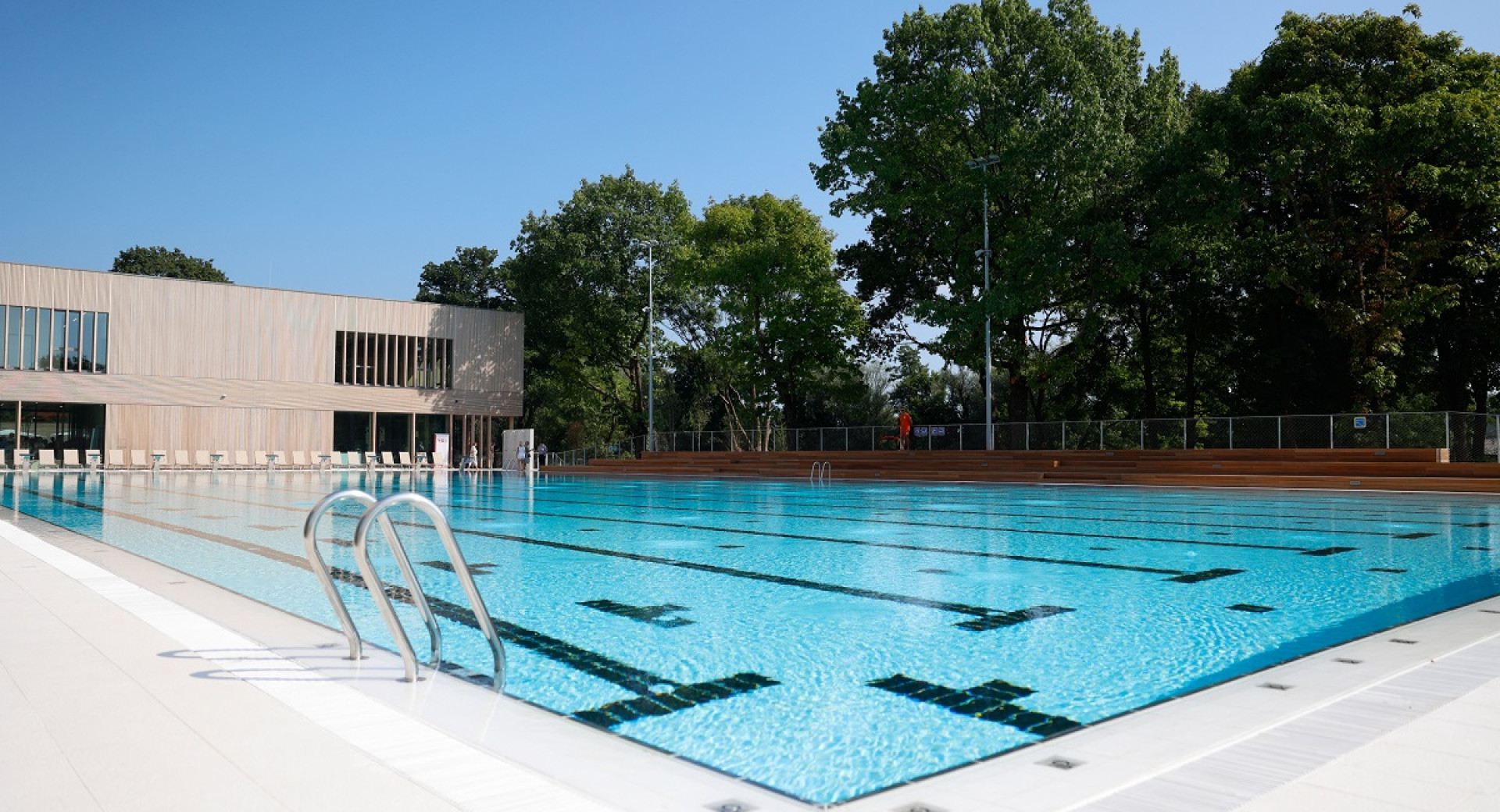 A view of the large outdoor pool surrounded by greenery and a wooden building.