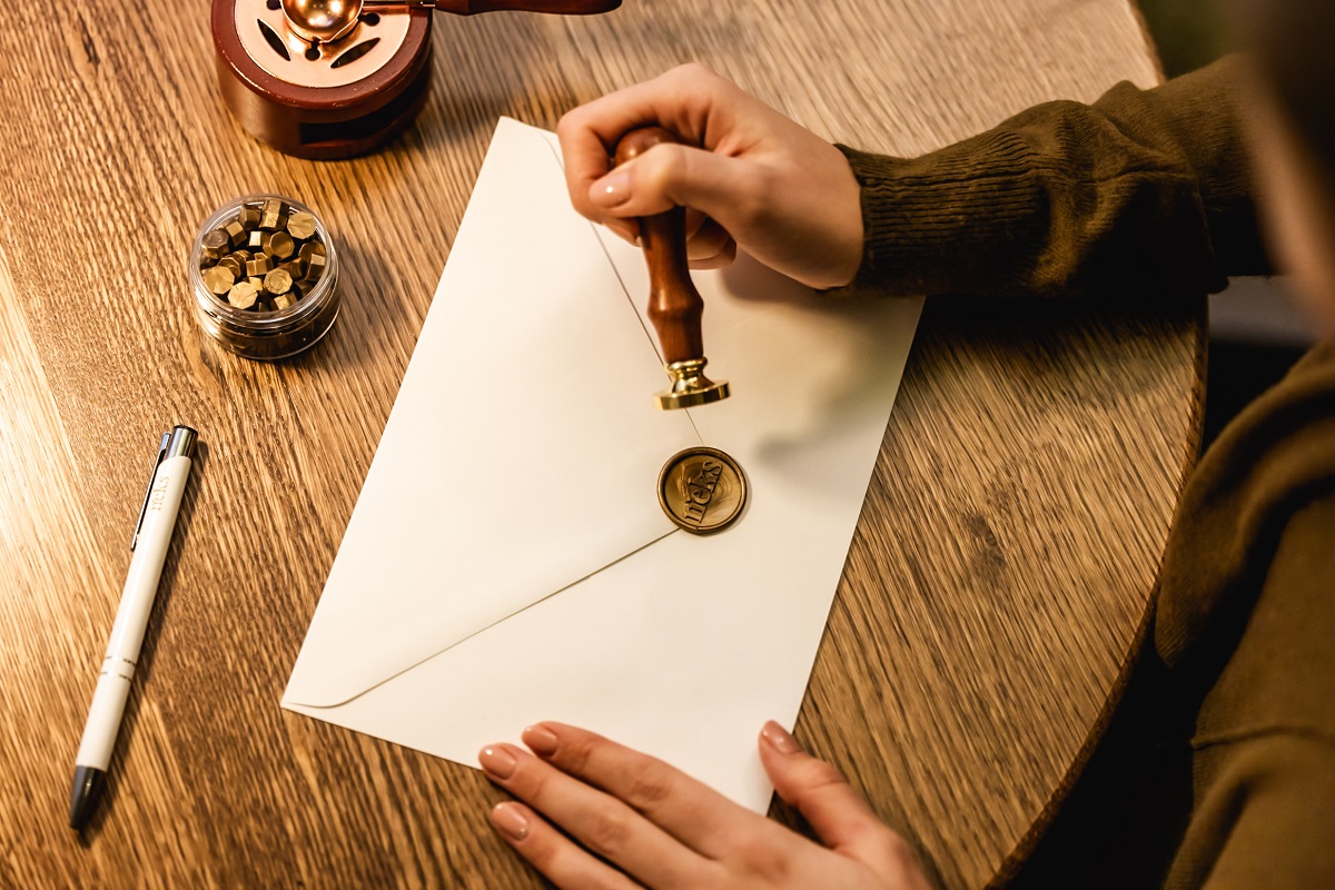 A woman's hand on a wooden table seals a white envelope with a wax seal.