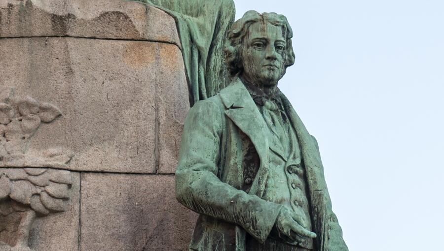 A bronze statue of a man in formal clothing, holding a book in one hand and gesturing with the other. The figure appears to be standing on a stone pedestal adorned with decorative carvings.