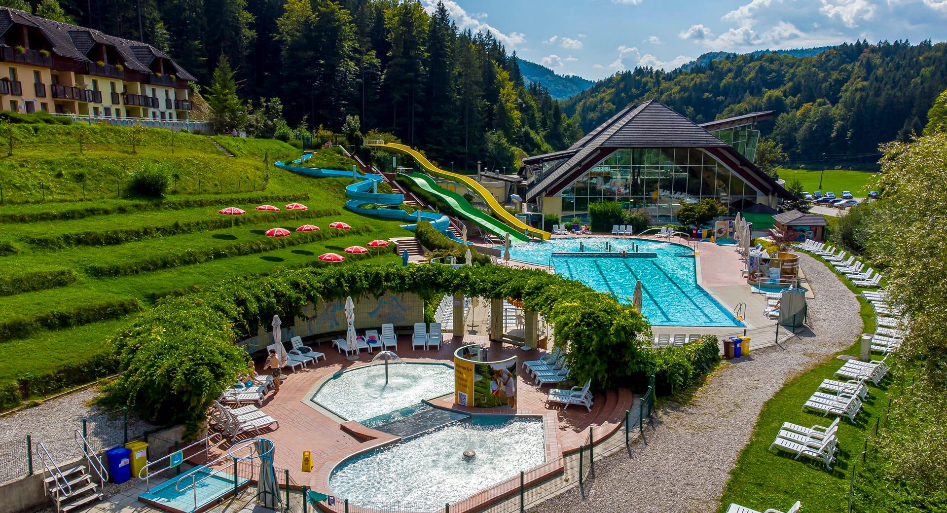View of the outdoor pool complex set in lush nature with several pools, water slides, sun loungers, and surrounding wooded hills.