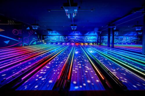 A neon-lit bowling alley with multiple parallel lanes highlighted by colourful LED lights and blue ambient lighting.